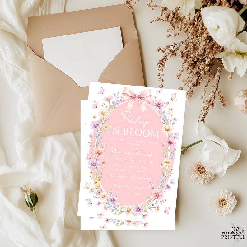 May include: A baby shower invitation with a pink oval center featuring the text "Baby in Bloom." The invitation is surrounded by a floral wreath with butterflies. A light brown envelope and a blank white card are visible in the background.