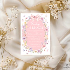 May include: A baby shower invitation with the text "Baby in Bloom" in a pink oval, surrounded by a floral border with butterflies. The invitation includes event details and is set against a cream-colored fabric.