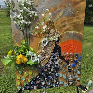 May include: A mixed-media canvas art piece featuring a silhouette of a person with long twine hair, wearing a black dress adorned with colorful rhinestones. The figure holds a miniature lantern amidst glass marbles, set against a gold and orange sunset.