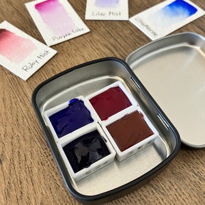 May include: A metal watercolor paint tin with four pans of paint. The pans are filled with blue, dark blue, red, and brown paint. The tin is open and the lid is off to the side. There are four cards with watercolor swatches and names: Ruby Mist, Purple Lake, Lilac Mist, and Ultramarine.