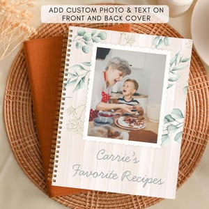Op de afbeelding: Een spiraalgebonden receptenboek met een aangepaste foto en tekst op de omslag. De omslag toont een foto van een vrouw en een kind, met de tekst "Carrie's Favorite Recipes". Het boek ligt op een geweven placemat.