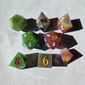 May include: A collection of colorful polyhedral dice for tabletop games. The dice are various shapes, including D4, D6, D8, D12, D20, and D100. Colors include green, red, and clear with gold or white numbering. A single brown tile with the number 6 is also present.