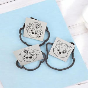 Puede incluir: Tres cortadores de galletas grises oscuros con forma de caras de perros de dibujos animados, cada uno con una imagen cuadrada de un personaje de perro diferente. Las imágenes son dibujos lineales en blanco y negro. Los cortadores están sobre una superficie azul claro.