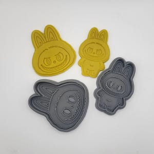 May include: Four cookie cutters in the shape of a cartoon bunny with a sinister grin. Two are yellow, and two are gray. The bunny has large ears and a round body. The cutters are likely for making cookies or other baked goods.