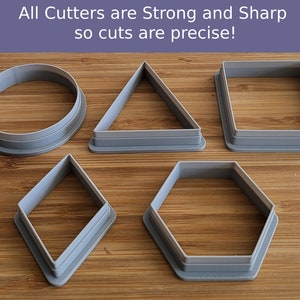 May include: Six different cookie cutters made of gray plastic. The cutters are shaped like a circle, triangle, square, diamond, hexagon, and a circle with a raised edge. The cutters are on a wooden surface.
