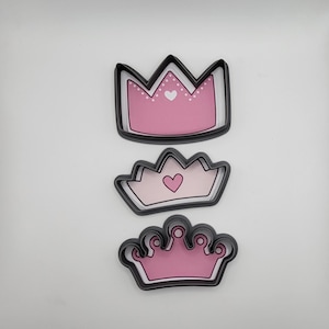 May include: Three pink and black crown-shaped cookie cutters. The largest cutter has a heart and white dots on the crown. The middle cutter has a heart on the crown. The smallest cutter has a simple crown design.