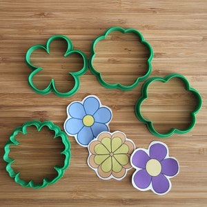 May include: A set of five cookie cutters in the shape of flowers. The cutters are green and feature different flower designs. There are also four paper flower cutouts in blue, yellow, and purple.