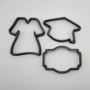 May include: Three metal cookie cutters in the shape of a graduation cap, a gown, and a blank rectangle.