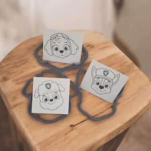 Puede incluir: Tres cortadores de galletas grises con forma de caras de perro, cada uno sosteniendo un cuadrado blanco con una cara de perro de dibujos animados diferente impresa. Los cortadores están sobre una superficie de madera.