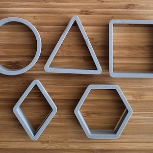 Peut inclure: Cinq emporte-pièces en plastique gris de formes différentes : cercle, triangle, carré, losange et hexagone. Les emporte-pièces sont sur une surface en bois.