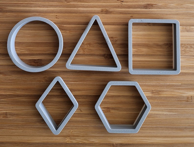 May include: Five gray plastic cookie cutters in different shapes: circle, triangle, square, diamond, and hexagon. The cutters are on a wooden surface.