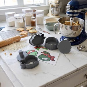 May include: A set of grey plastic cookie cutters shaped like cartoon turtle heads with red masks, accompanied by printed reference cards showing the character design, placed on a white marble countertop near a blue stand mixer and baked cookies.