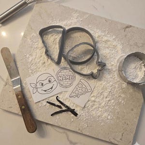 May include: Two grey plastic cookie cutters, one shaped like a pizza slice and one like a turtle head, alongside three paper templates featuring a turtle face, a pizza slice, and a circular brick pattern with the text NAME, plus vanilla beans.