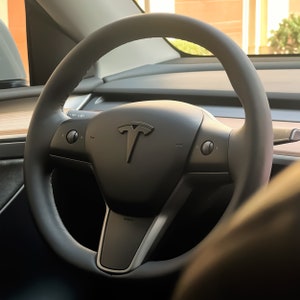 May include: A black leather steering wheel with a silver Tesla logo in the centre. The steering wheel has two buttons on each side.