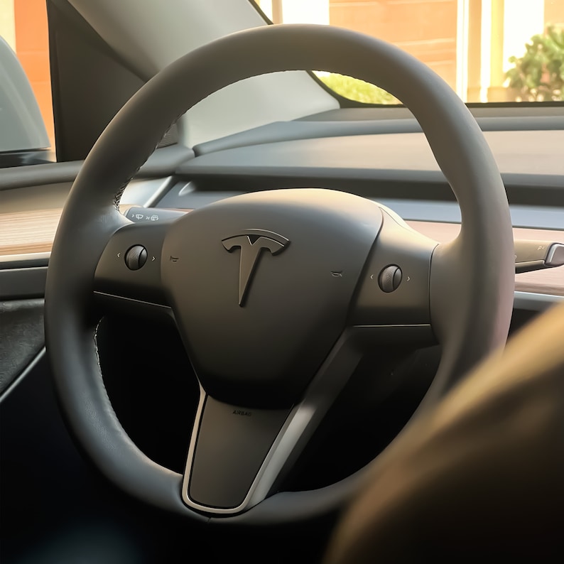 May include: A black leather steering wheel with a silver Tesla logo in the centre. The steering wheel has two buttons on each side.