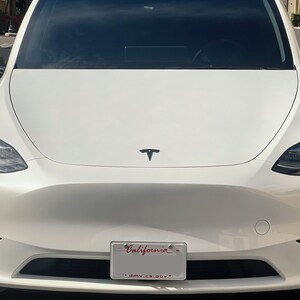 May include: White Tesla Model Y with a California number plate. The car has a black Tesla logo on the bonnet.