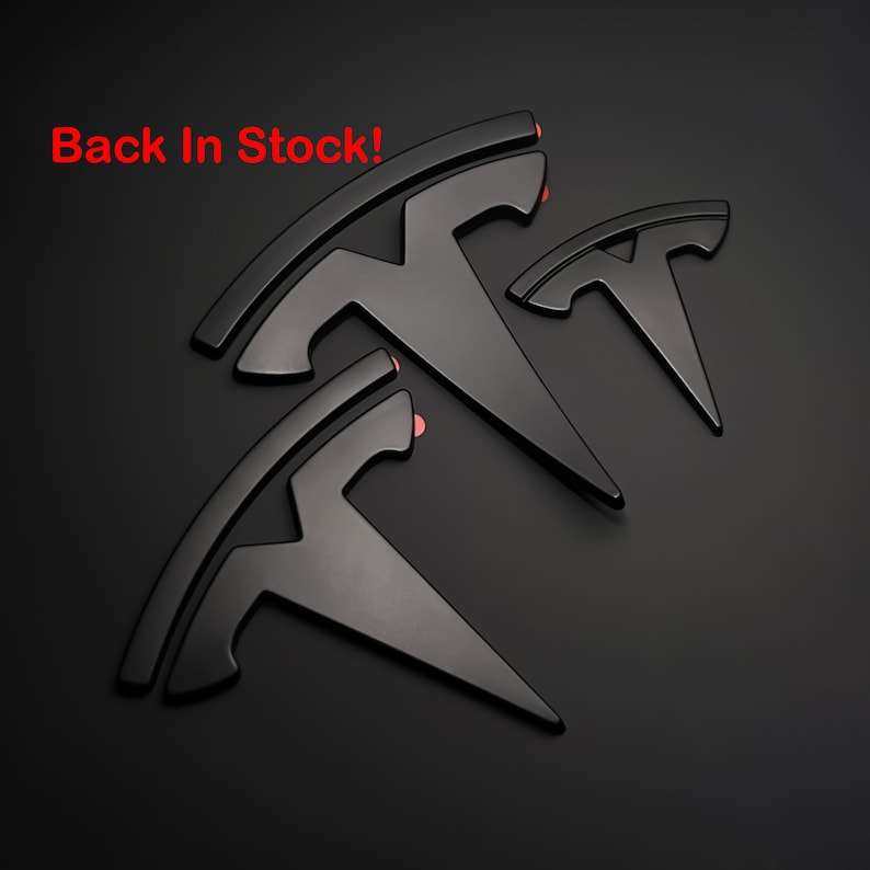 May include: Three black metal Tesla logos of different sizes, arranged in a staggered pattern on a black background. The largest logo is at the top, and the smallest logo is at the bottom. The text "Back In Stock!" is written in red at the top of the image.