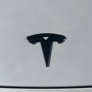 May include: A black Tesla logo on a white car. The logo is a stylised letter T with a curved line above it.