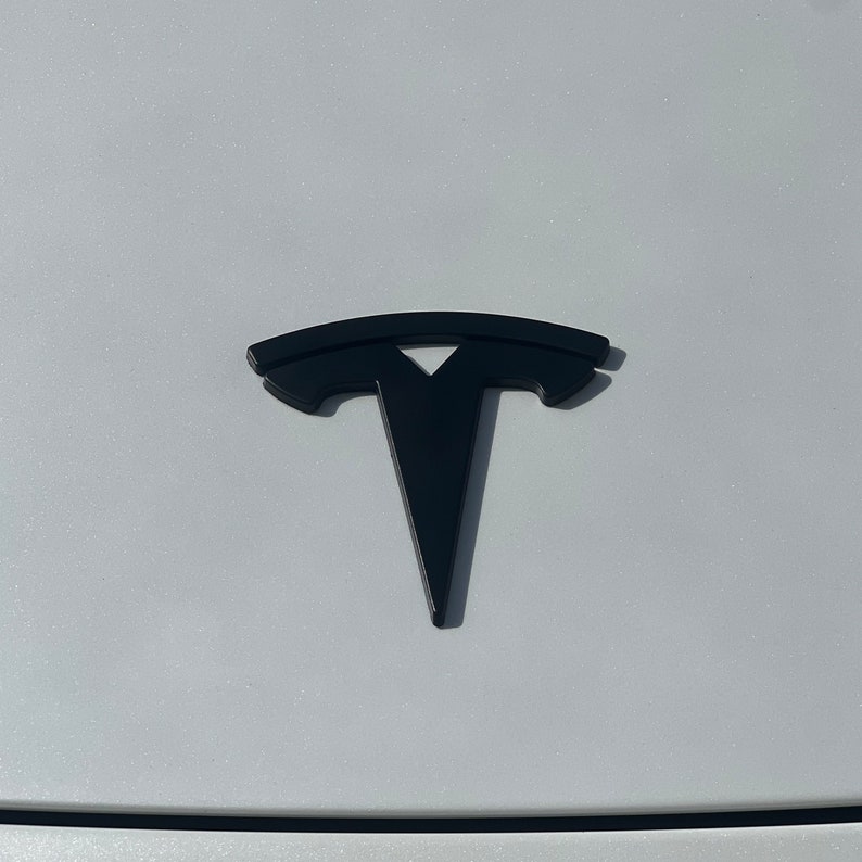 May include: A black Tesla logo on a white car. The logo is a stylised letter T with a curved line above it.