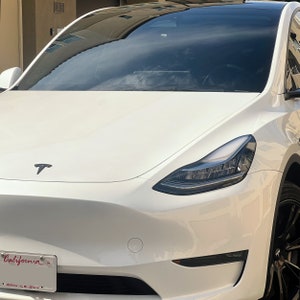 May include: White Tesla Model Y with black headlights and a black Tesla logo on the bonnet. The car has a California number plate.