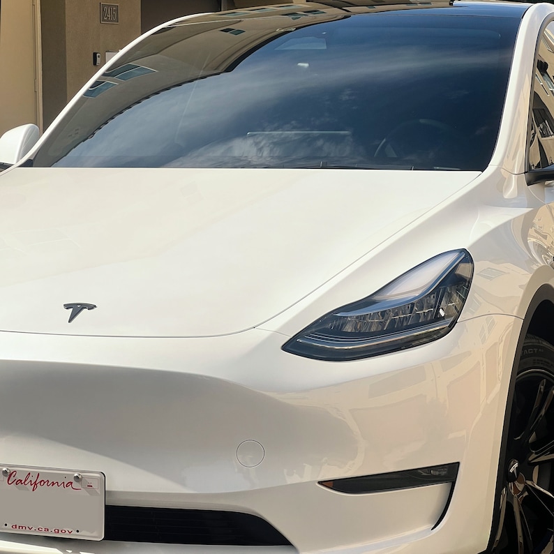 May include: White Tesla Model Y with black headlights and a black Tesla logo on the bonnet. The car has a California number plate.