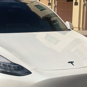 May include: A white Tesla Model Y with a black Tesla logo on the bonnet. The car is parked in front of a building with two garage doors.