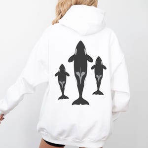May include: A white hooded sweatshirt featuring a graphic print on the back. The design displays three stylised black orca whales arranged vertically, with the central whale being the largest and two smaller whales positioned on either side.