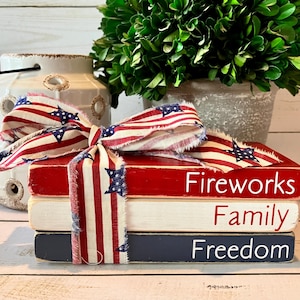 May include: Three wooden blocks stacked on top of each other. The top block is red and says "Fireworks". The middle block is white and says "Family". The bottom block is blue and says "Freedom". A red, white, and blue ribbon is tied around the blocks.