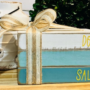 May include: Three wooden blocks stacked on top of each other. The top block is white with the words "Don't Be" in yellow lettering. The middle block is blue with the word "Salty" in yellow lettering. The bottom block is blue. The blocks are tied together with a brown and white ribbon.