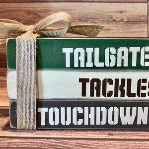 May include: A stack of three wooden blocks with the words "Tailgates", "Tackles", and "Touchdowns" painted on them. The blocks are tied together with twine and a brown bow.