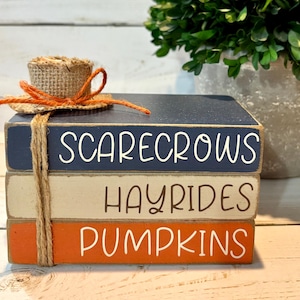 May include: Stacked wooden blocks painted in navy, beige, and orange, with the words "SCARECROWS," "HAYRIDES," and "PUMPKINS" in white. A small straw hat with an orange ribbon sits on top, tied with twine. A decorative fall-themed item.