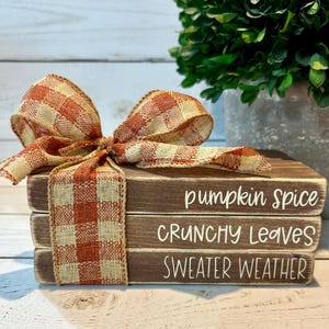 May include: Three wooden blocks stacked together, each with a white script phrase: "pumpkin spice", "crunchy leaves", and "sweater weather". An orange and cream plaid ribbon adorns the blocks. The blocks are brown with a distressed finish, creating a rustic aesthetic.