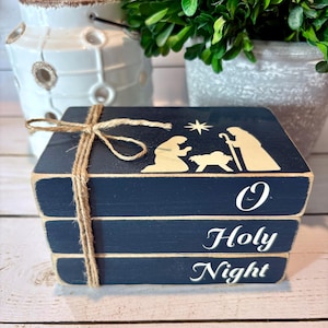 May include: A navy blue wooden block set tied with twine. The top block features a nativity scene silhouette and the words "O Holy Night" in white script. Decorative Christmas decor.