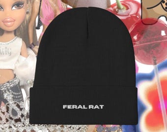 May include: Black beanie with the text "FERAL RAT" embroidered in white. The hat is a solid color and has a folded cuff. The background includes images of dolls and candy.