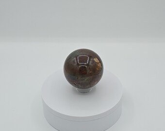 May include: A polished sphere of iridescent, multi-colored jasper, with brown, red, and green hues, displayed on a white pedestal.