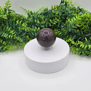 May include: A dark purple and green gemstone sphere on a white circular display stand. The sphere is smooth and polished.