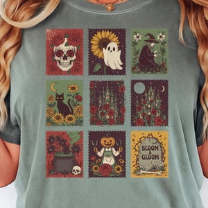 May include: Moss green t-shirt featuring nine postage stamp-style graphics with Halloween themes. The graphics include a skull, ghost, witch, black cat, castle, pumpkin, and the words "Bloom & Gloom".