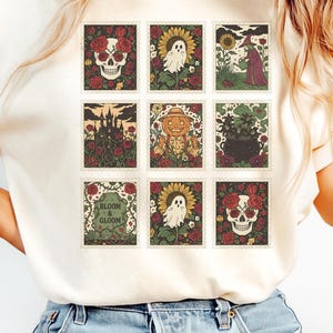 May include: Ivory-colored t-shirt featuring a grid of Halloween-themed postage stamp designs. The stamps showcase images of skulls, ghosts, sunflowers, a witch, a haunted castle, a scarecrow, a cauldron, and the text "Bloom & Gloom".