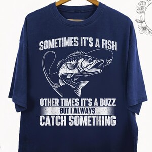 May include: Navy blue t-shirt with a fishing-themed graphic. The design features a fish and fishing rod, along with the text "SOMETIMES IT'S A FISH, OTHER TIMES IT'S A BUZZ BUT I ALWAYS CATCH SOMETHING". The shirt is hanging on a wooden hanger.