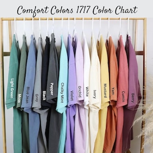May include: A color chart of Comfort Colors 1717 t-shirts, showcasing a variety of hues. The shirts are displayed on hangers, with colors including light green, moss, blue jean, gray, black, pepper, chalky mint, violet, orchid, white, ivory, mustard, yam, cumin, and berry.