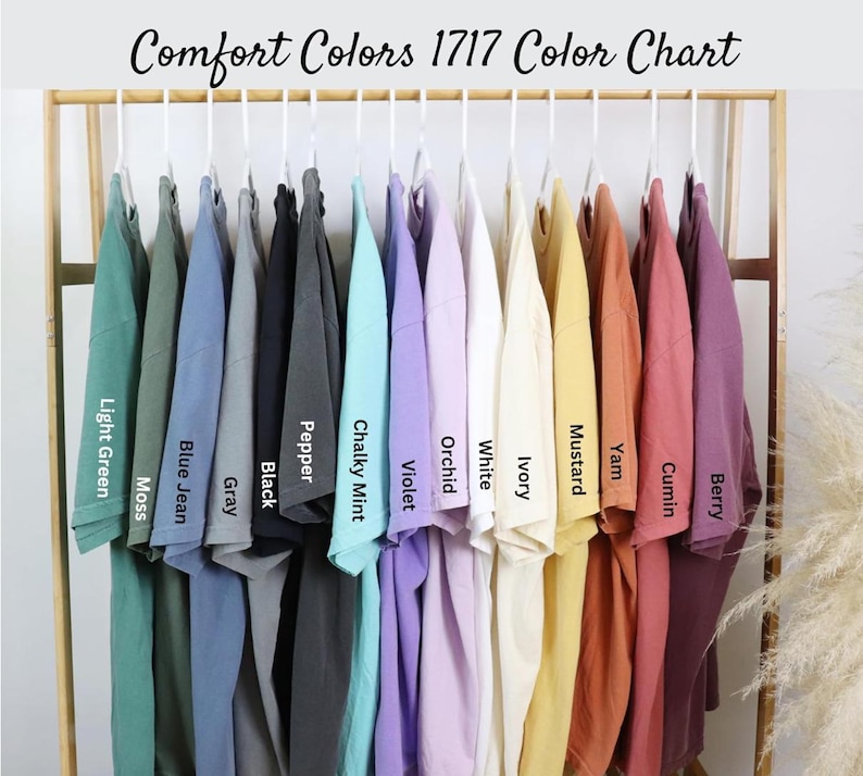 May include: A color chart of Comfort Colors 1717 t-shirts, showcasing a variety of hues. The shirts are displayed on hangers, with colors including light green, moss, blue jean, gray, black, pepper, chalky mint, violet, orchid, white, ivory, mustard, yam, cumin, and berry.