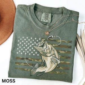 May include: A moss green t-shirt with a graphic of a fish jumping for a lure, set against a camouflage and American flag design. The shirt has a crew neck and short sleeves. A silver necklace with a small pendant is on the shirt. The word "MOSS" is at the bottom.