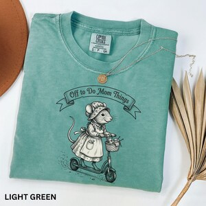 May include: Light green t-shirt with the text "Off to Do Mom Things" above an illustration of a mouse riding a scooter. The mouse wears a bonnet and apron and carries a basket. The shirt has a crew neck and short sleeves.