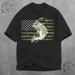 May include: Black t-shirt featuring a graphic of a leaping fish with a fishing hook, set against a camouflage and American flag design. The fish is light beige with dark green accents. The shirt has a classic crew neck.