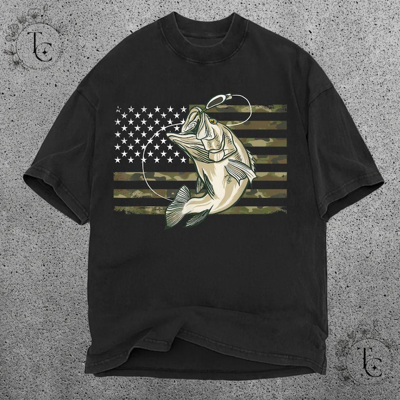 May include: Black t-shirt featuring a graphic of a leaping fish with a fishing hook, set against a camouflage and American flag design. The fish is light beige with dark green accents. The shirt has a classic crew neck.