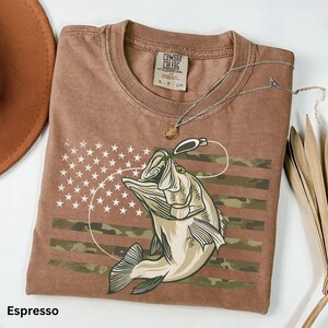 May include: Espresso-colored t-shirt featuring a graphic of a fish leaping towards a lure, set against a camouflage-striped and star-studded background. The shirt includes a Comfort Colors tag and a silver necklace.