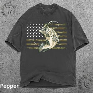 May include: Dark gray t-shirt featuring a graphic of a fish jumping out of the water with a fishing hook. The design incorporates the American flag with a camouflage pattern. The word "Pepper" is at the bottom.