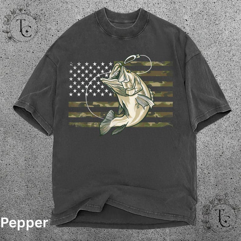 May include: Dark gray t-shirt featuring a graphic of a fish jumping out of the water with a fishing hook. The design incorporates the American flag with a camouflage pattern. The word "Pepper" is at the bottom.