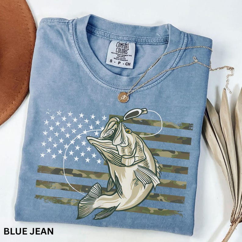 May include: Blue jean colored t-shirt with a graphic of a fish jumping for a lure, set against a camouflage and star design. The shirt has a crew neck and short sleeves. A gold necklace with a pendant is draped across the shirt.