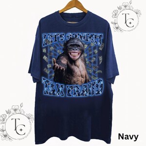 May include: Navy blue t-shirt with a graphic of a smiling chimpanzee and the text "LET'S COMMIT TAX FRAUD" in a blue, pixelated font. The design includes falling dollar bills, creating a humorous and eye-catching graphic.